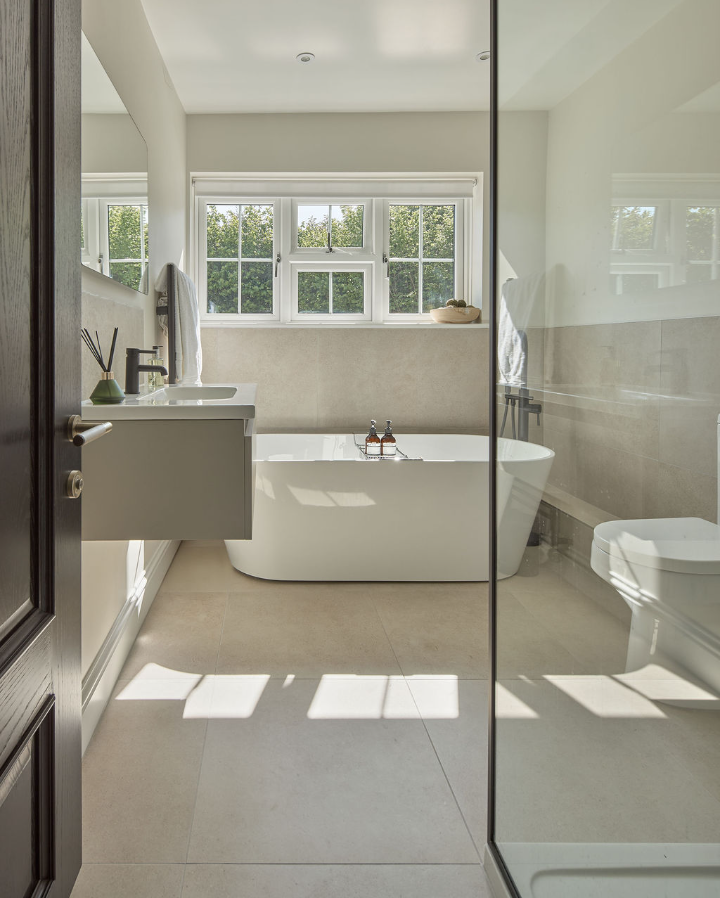 Modern bathroom with a freestanding white bathtub below a large window, floating sink on the left, and a glass shower on the right. Neutral tones and natural light create a bright, minimalistic atmosphere.