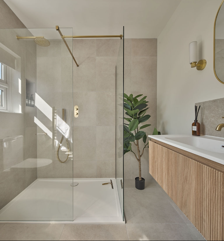 Modern bathroom with a walk-in glass shower featuring gold fixtures, a floating wooden vanity with a white sink, a potted plant, and neutral-toned walls and flooring. Natural light streams in through a window.
