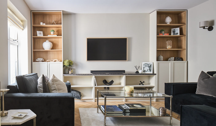 Modern living room with two dark sofas, a glass coffee table, built-in shelves with decor items, and a wall-mounted TV above a long console. Natural light comes from a window on the left.