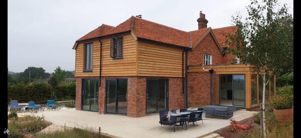 A modern two-story house with a mix of wooden siding and red brick exterior, large glass doors, a patio area with outdoor seating, and a partially visible garden.