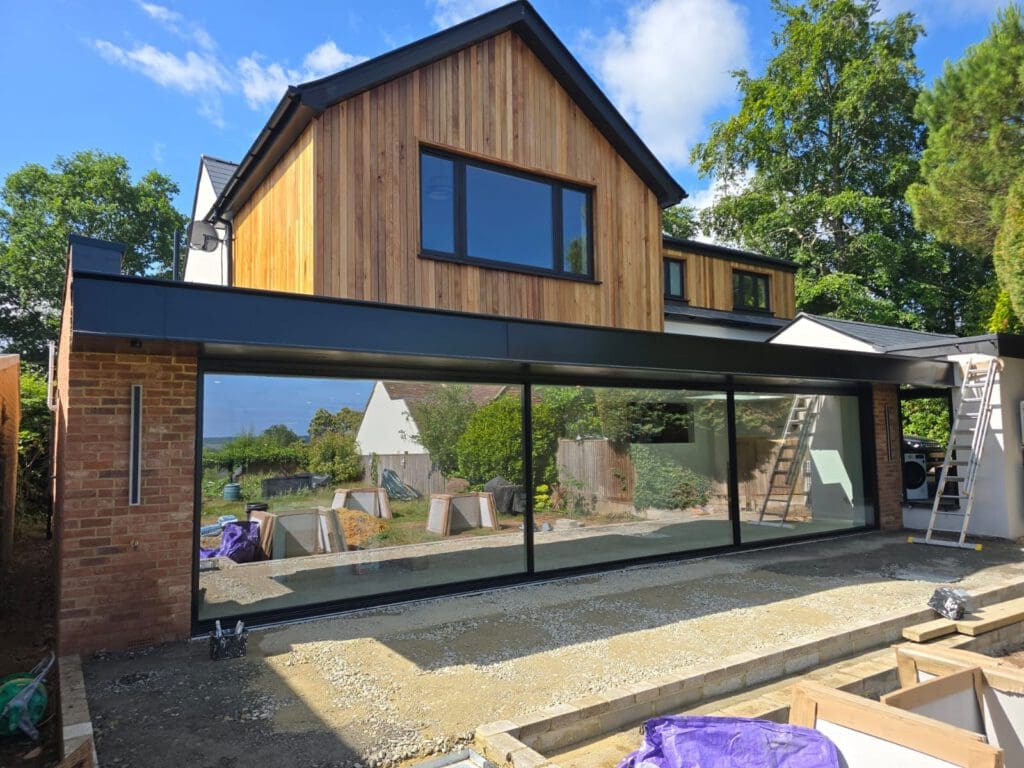 A modern two-story house with wood and brick exterior, large floor-to-ceiling glass windows, and a partially completed patio area, surrounded by greenery and construction materials.