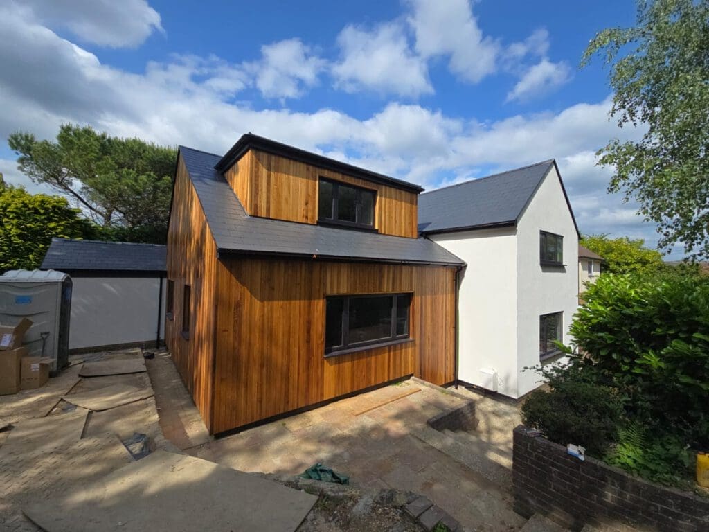 A modern house extension with vertical wooden cladding and large windows is attached to a white, two-story home. The sky is partly cloudy, and there are trees and greenery around the building.