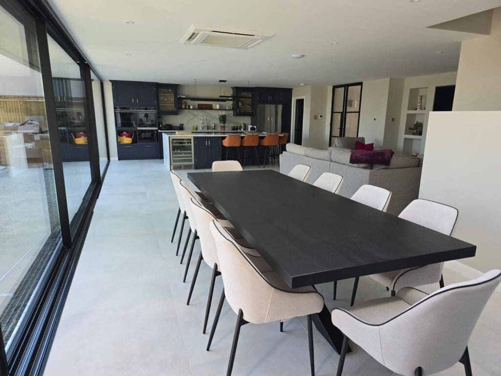 Modern open-plan kitchen and dining area with a long black table, cream chairs, dark cabinets, orange bar stools, and large glass windows overlooking an outdoor space.