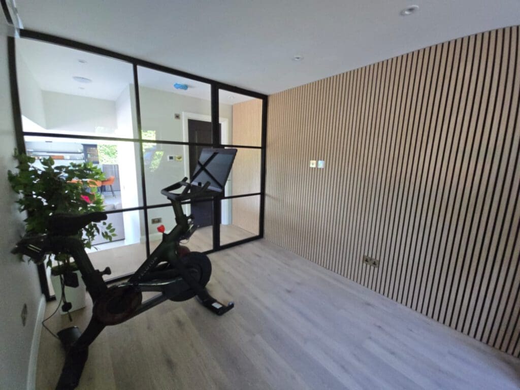 A modern home gym with a stationary bike, light wood flooring, slatted wood wall, and a glass partition separating the room from the rest of the house; plants sit beside the bike.