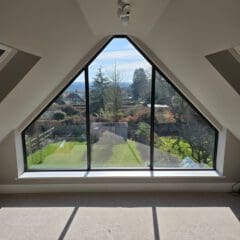 A triangular floor-to-ceiling window offers a clear view of a sunny yard with trees, a driveway, and distant hills. Sunlight streams into the beige-carpeted room, casting sharp shadows on the floor.