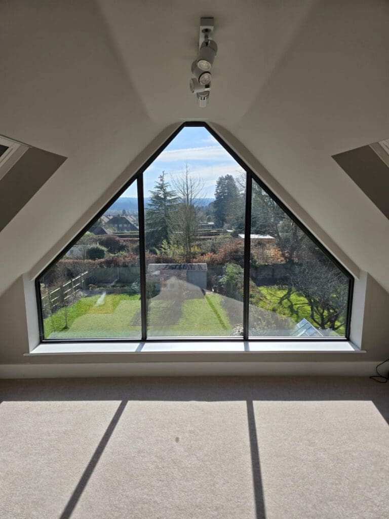 A triangular floor-to-ceiling window offers a clear view of a sunny yard with trees, a driveway, and distant hills. Sunlight streams into the beige-carpeted room, casting sharp shadows on the floor.