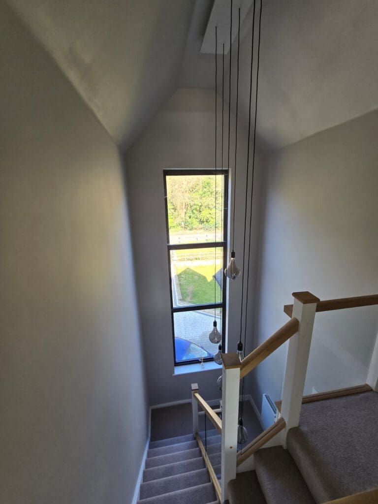 A modern staircase with a grey carpet, wooden handrail, and hanging pendant lights, leading down toward a tall window that overlooks a garden with greenery outside.