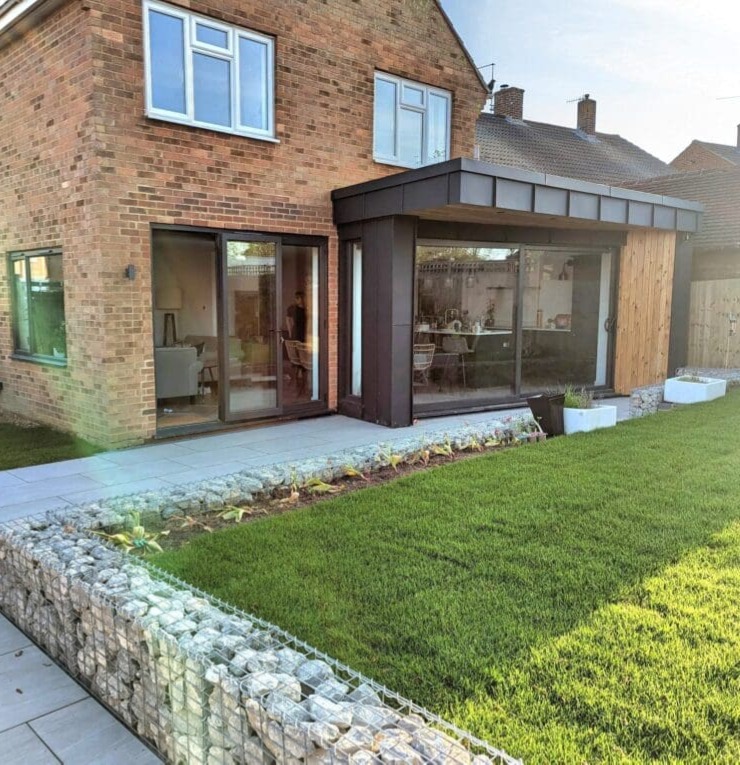 A modern brick house with large glass sliding doors, an extension with a dark frame, a stone-bordered flower bed, and a neatly mowed lawn in the foreground.