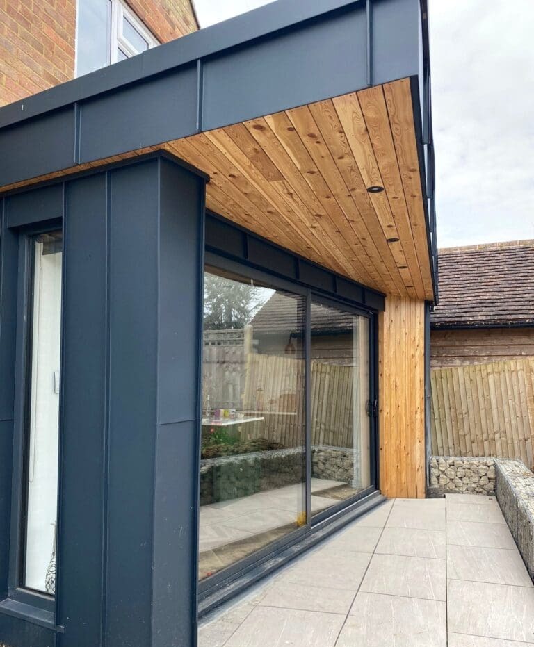 Modern house extension with black metal siding, wood-paneled ceiling, and large sliding glass doors opening onto a tiled patio. Wooden fencing and stone gabion walls enclose the outdoor area.