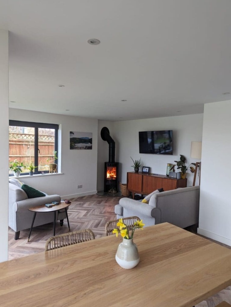Modern living room with light gray sofas, a wooden coffee table, a lit wood-burning stove, a TV mounted on the wall, indoor plants, and a dining table with a vase of yellow flowers in the foreground.