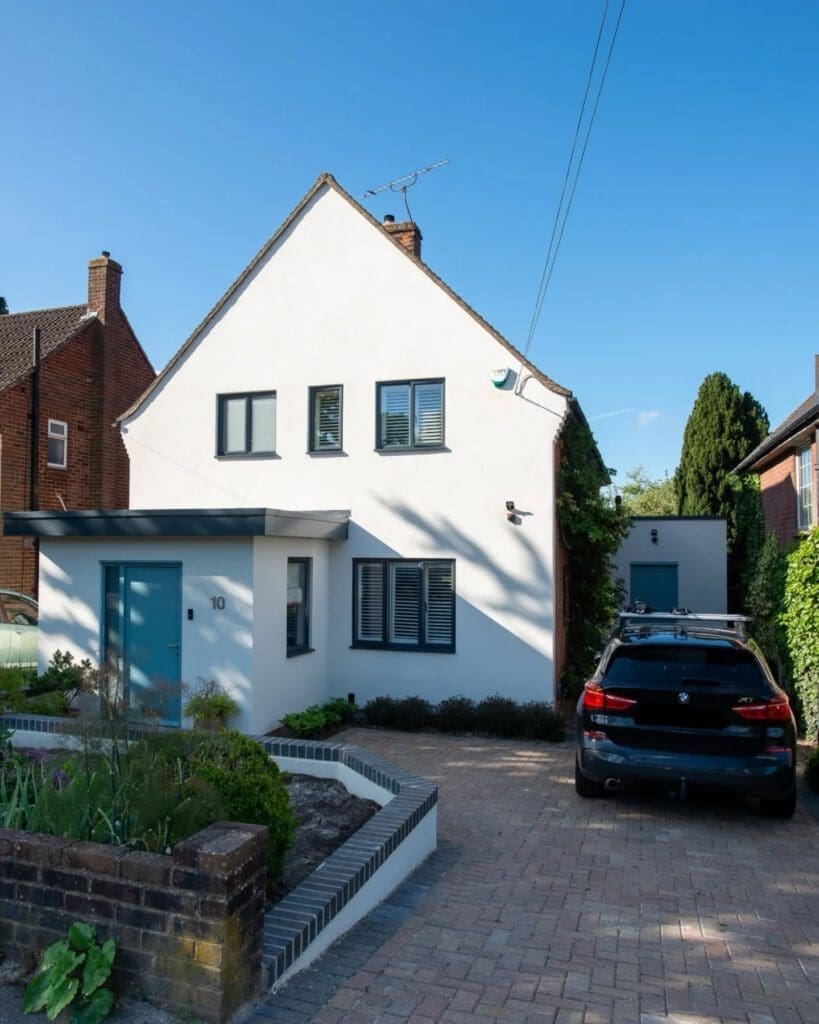 Modern white two-story house with blue door and window frames, a sloped roof, and a brick driveway. A black car is parked in the driveway, and there are plants and shrubs along the front garden.