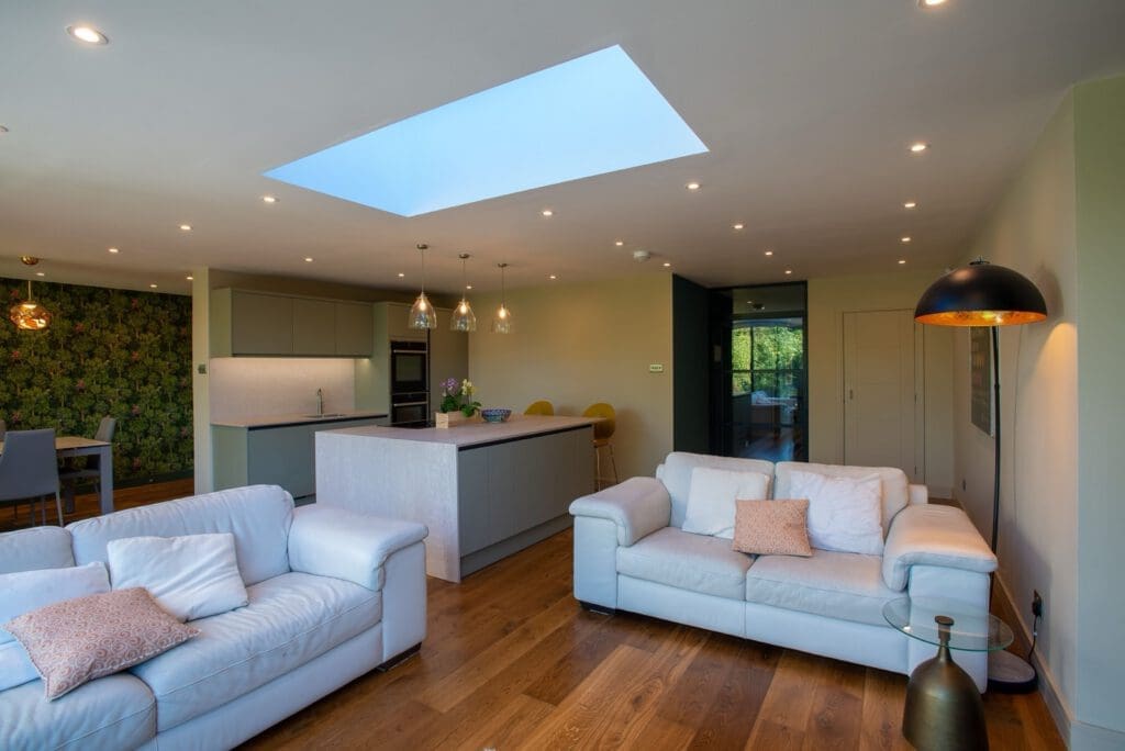 Modern open-plan living area with white sofas, wooden floor, large kitchen island, pendant lights, a skylight, and green floral accent wall; space is bright and airy with contemporary decor.