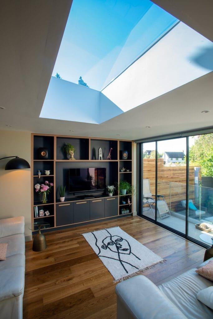 A modern living room with wooden floors, a large skylight, built-in shelves around a TV, white sofas, a line-art rug, and glass doors leading to a patio with greenery outside.