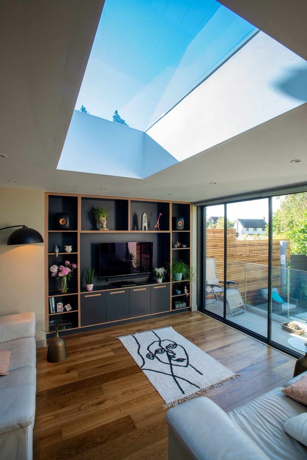 A modern living room with wooden floors, a large skylight, built-in shelves around a TV, white sofas, a line-art rug, and glass doors leading to a patio with greenery outside.