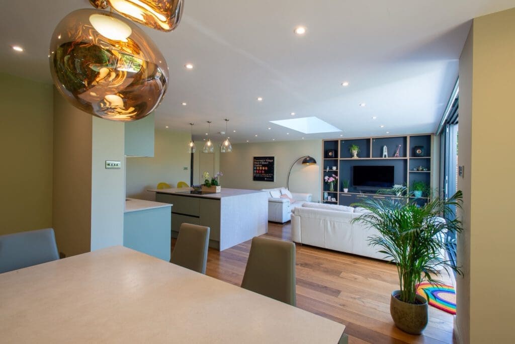Open-plan modern living space with dining table, kitchen island, and living area. Wooden floors, green plants, pendant lights, white sofas, built-in shelves, and large windows create a bright, cozy atmosphere.