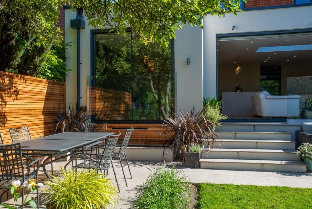 Modern backyard with a patio dining set, plants, and steps leading up to a contemporary house with large glass doors and windows that open to a bright living room with white furniture.