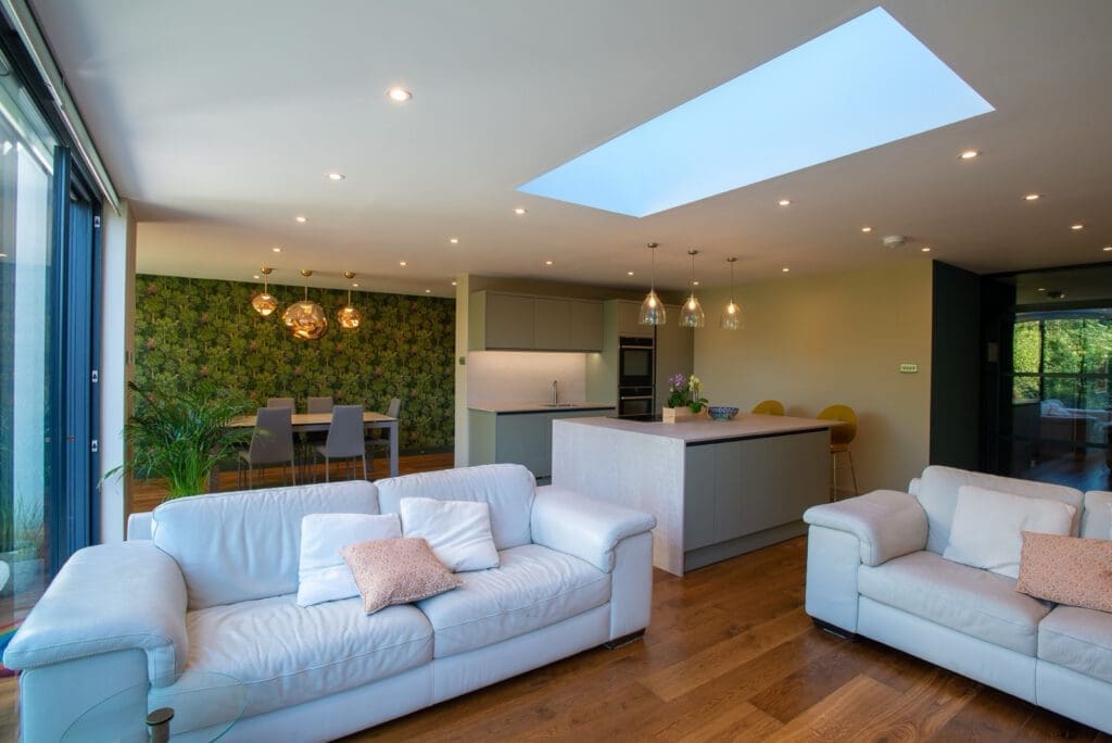 Open-plan living space with white sofas, a kitchen island with barstools, a skylight, wooden floors, and a dining area featuring a green floral accent wall and modern pendant lights. Large windows let in natural light.