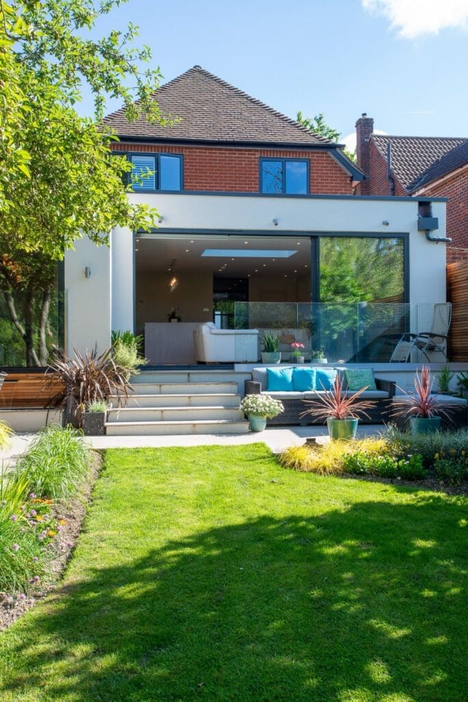 Modern house with large glass doors opening to a patio with outdoor seating, steps leading to a lush green lawn, and landscaped garden beds with plants and trees on a sunny day.