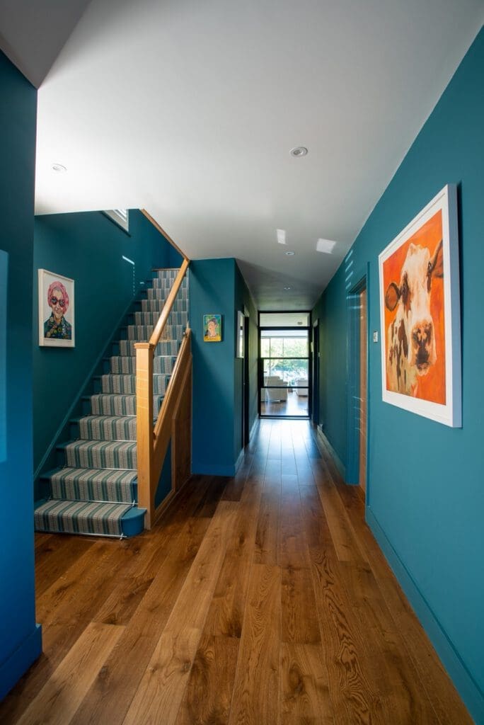 Bright hallway with teal walls, wood floor, and a staircase with striped carpet runner. Colorful artwork, including a cow portrait, decorates the walls. Natural light enters from glass doors at the end.