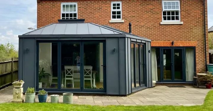 Modern glass and metal conservatory attached to a red-brick house, with large sliding doors, potted plants along the patio, and a green lawn in the foreground.