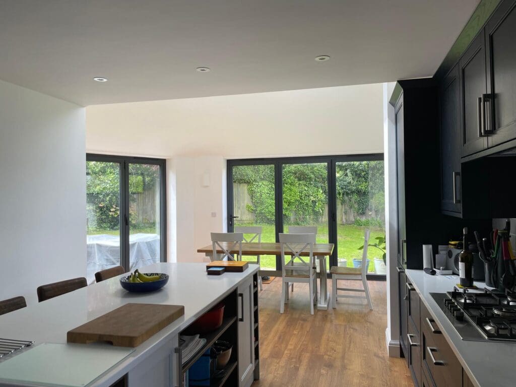 Modern kitchen with a central island, stools, and a view of a dining table near large floor-to-ceiling windows overlooking a green garden. Natural light fills the space, highlighting the wooden floor and dark cabinetry.