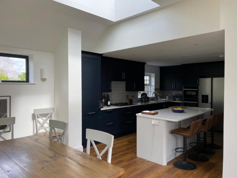 Modern kitchen with dark blue cabinets, white countertops, a large island with stools, and wooden flooring. A wooden dining table with white chairs is in the foreground. Natural light enters through a skylight and windows.