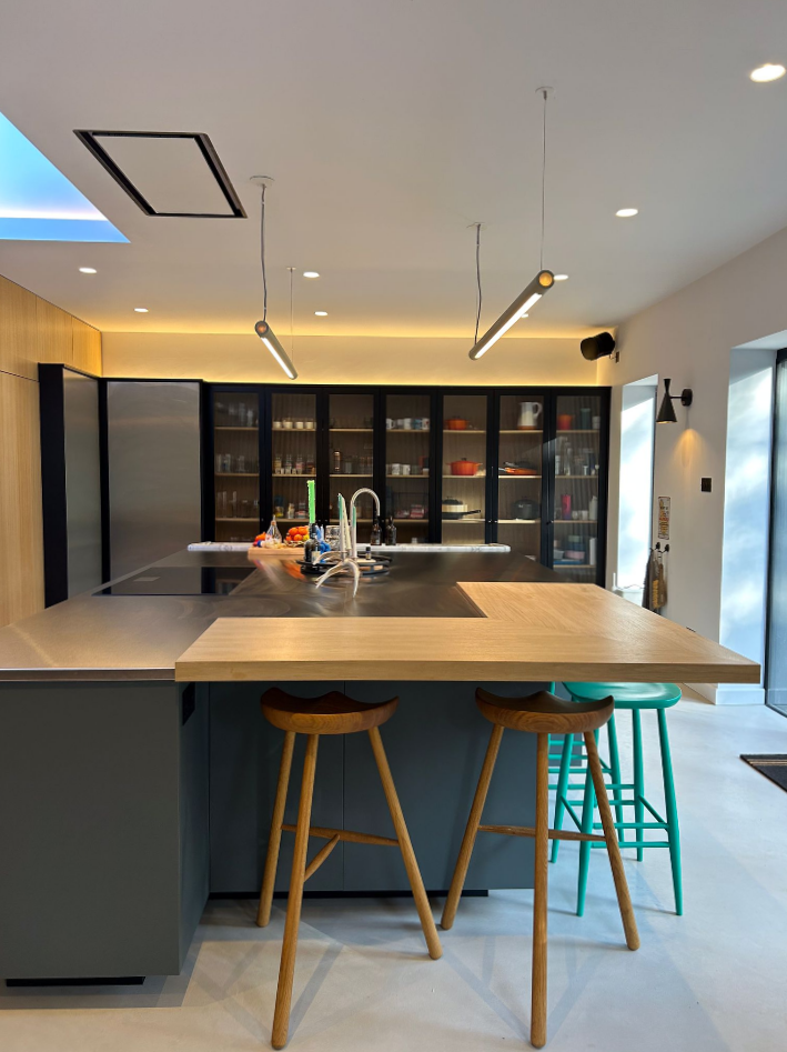 Modern kitchen with a large island, two wooden stools, and one green stool. The island has a sink and minimalist decor. Cabinets with glass doors line the back wall, and pendant lights hang from the ceiling.