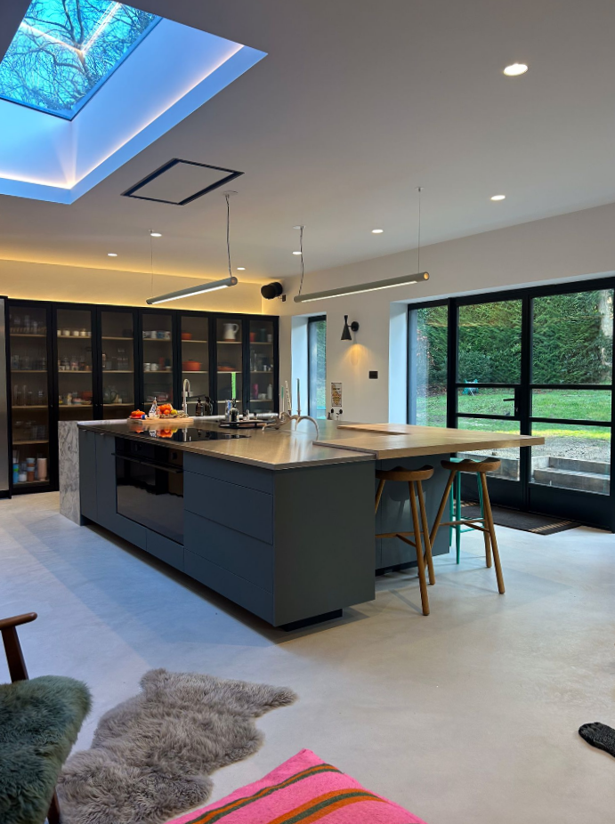 Modern kitchen with a large island, bar stools, skylight, and glass doors opening to a green yard. Shelves with dishes line the back wall, and a colorful rug and furry mat are on the light floor.