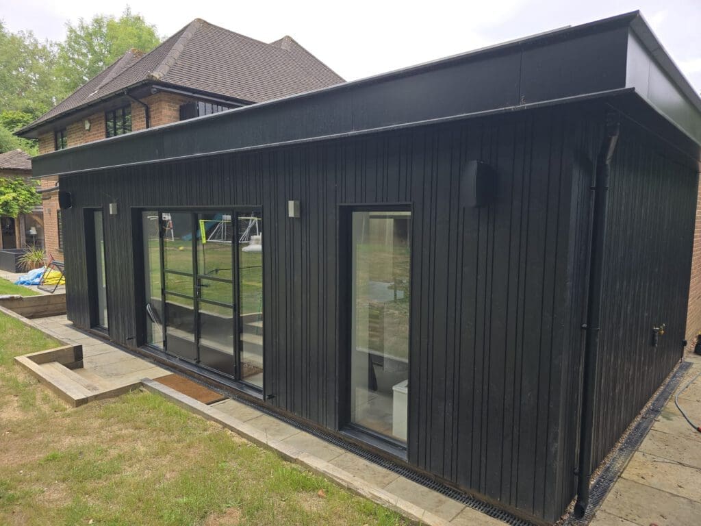 A modern black outbuilding with large glass doors and vertical siding sits on a stone patio in a grassy backyard, near a brick house.