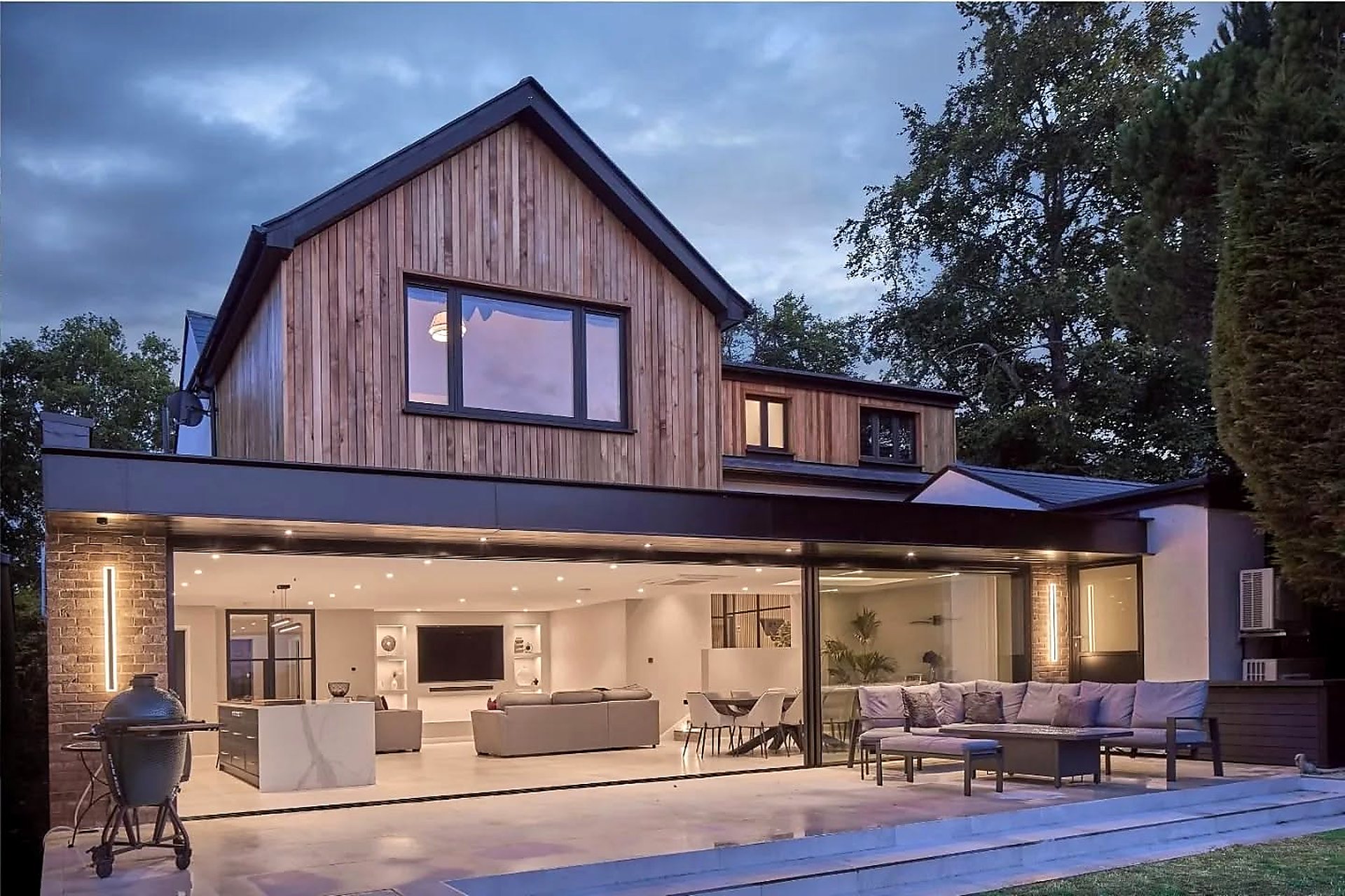 Modern two-story house with wood and brick exterior, large windows, and fully open sliding glass doors revealing a spacious, well-lit living area and outdoor patio with seating at dusk.
