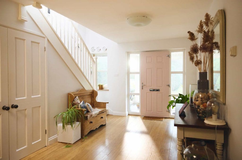 A bright hallway with wooden floors, a pink front door, a wooden bench with cushions and plants, a staircase, and a console table decorated with glass jars and dried flowers. Natural light streams in from the window and door.