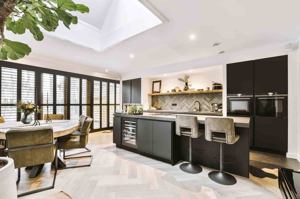 Modern kitchen with black cabinets, a large island with bar stools, built-in ovens, and open shelving. Adjacent dining area with round table and chairs, large windows with shutters, and a skylight above.