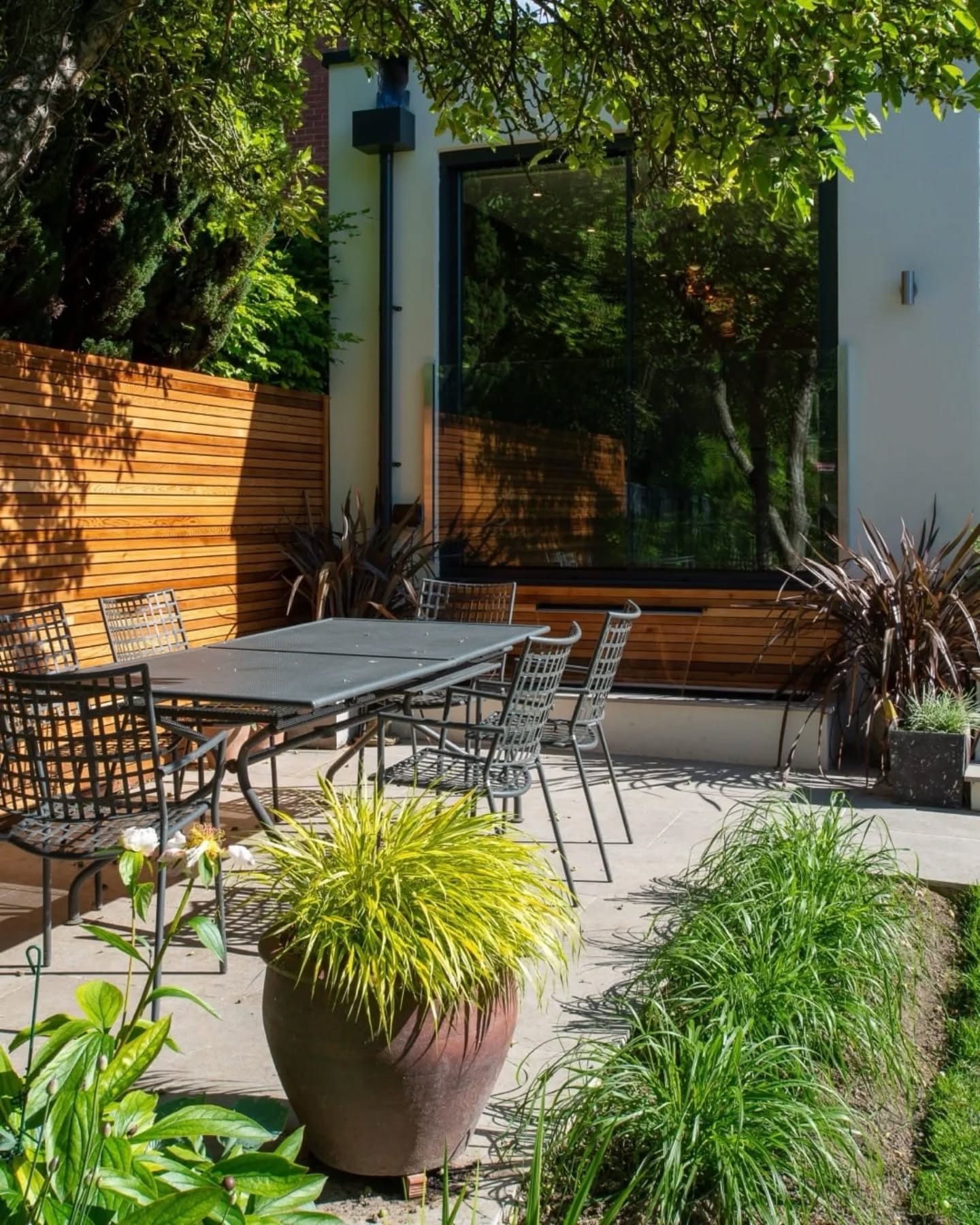 Modern patio with a black metal table and chairs, surrounded by greenery, wooden fence, and large window reflecting trees. Sunlight casts shadows on the concrete floor.