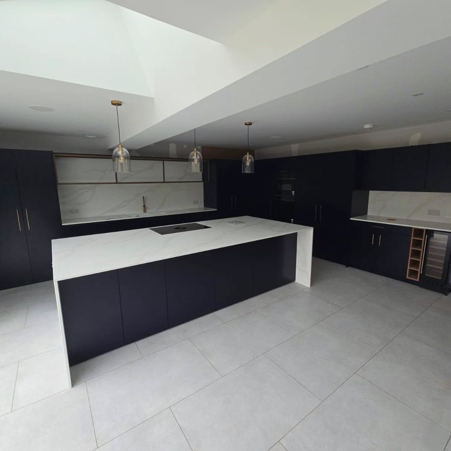 Modern kitchen with black cabinets, white countertops, a large island, pendant lights, and built-in appliances. The space features light gray tiled flooring and minimalist design.