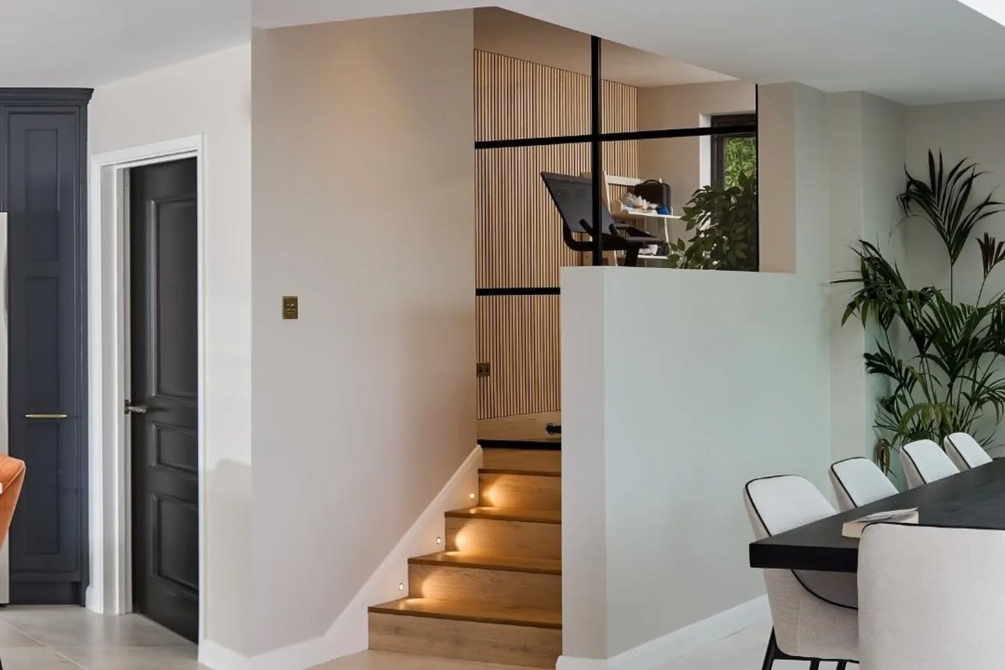 A modern home interior with a short staircase leading to a small office space with a desk and chair. The area features light wood steps with lights, white walls, and a dining table with chairs nearby.