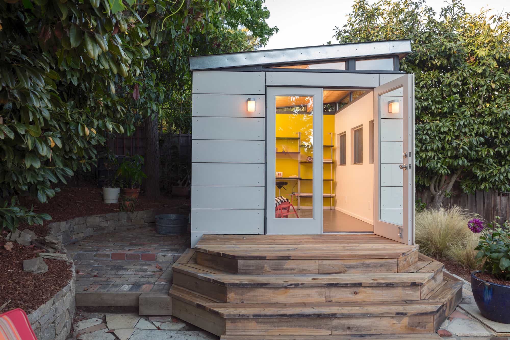 A small modern shed with white paneling, glass double doors, and wooden steps sits in a garden surrounded by trees and plants. Warm yellow light glows from inside, showing shelves and a chair.