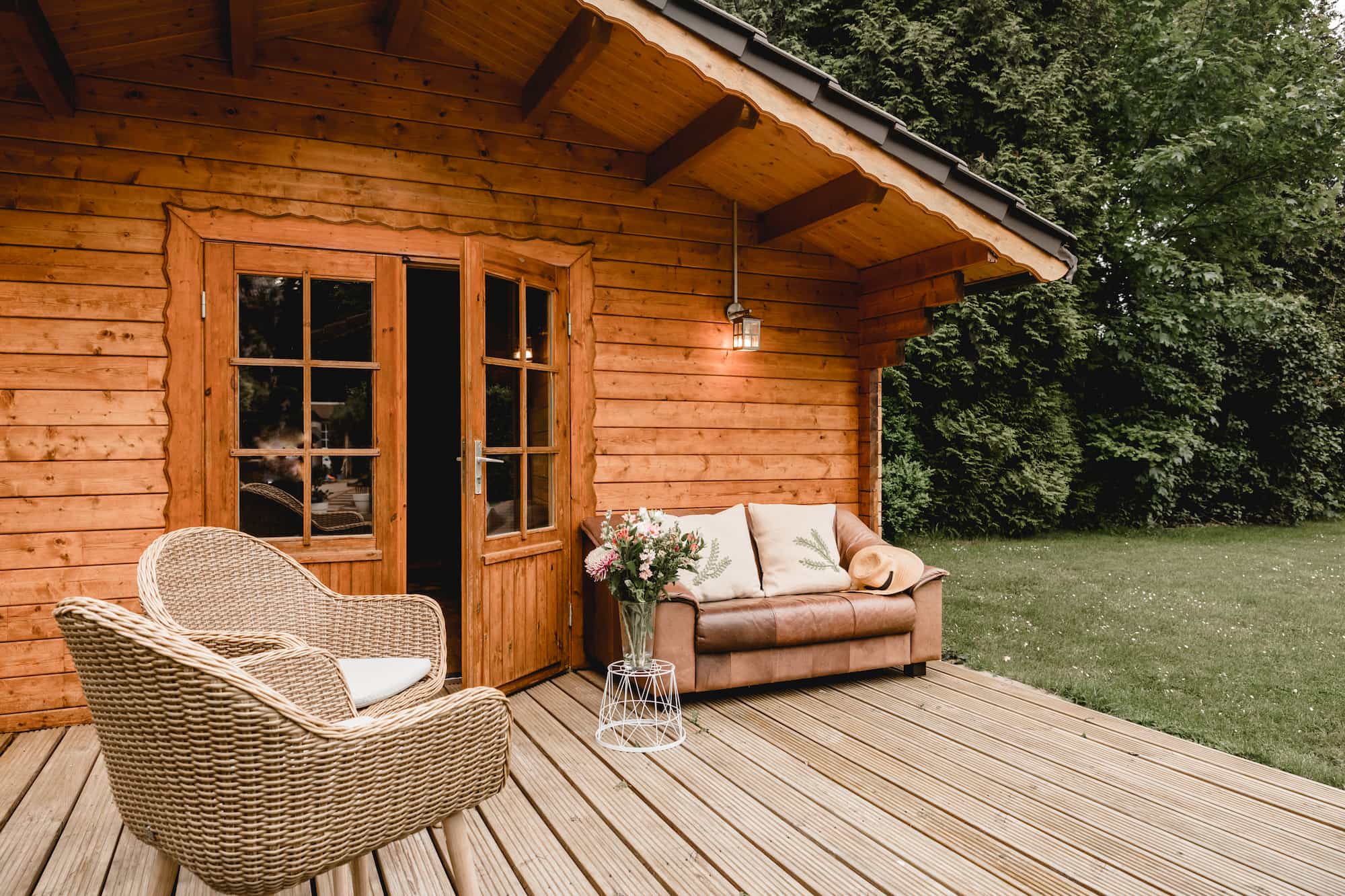 Wooden cabin with glass doors, a wicker chair, a brown couch with a blanket, flowers on a small table, and a green lawn with trees in the background.