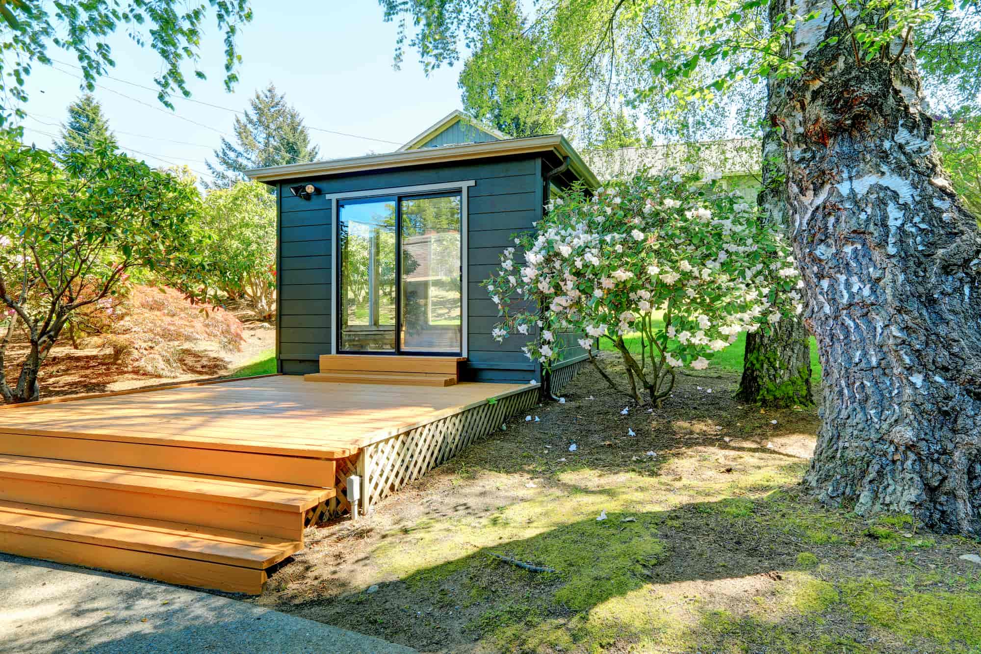 Small dark gray house with large glass doors, wooden steps leading to a deck, surrounded by green trees, blooming bushes, and a sunlit yard.